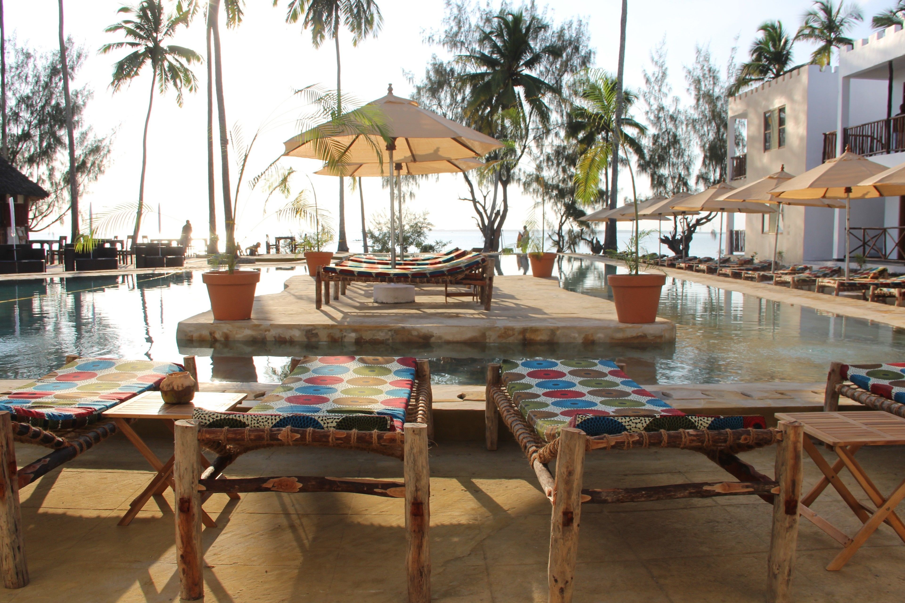 A poolside area with lounge chairs and umbrellas, surrounded by palm trees and a building with balconies.