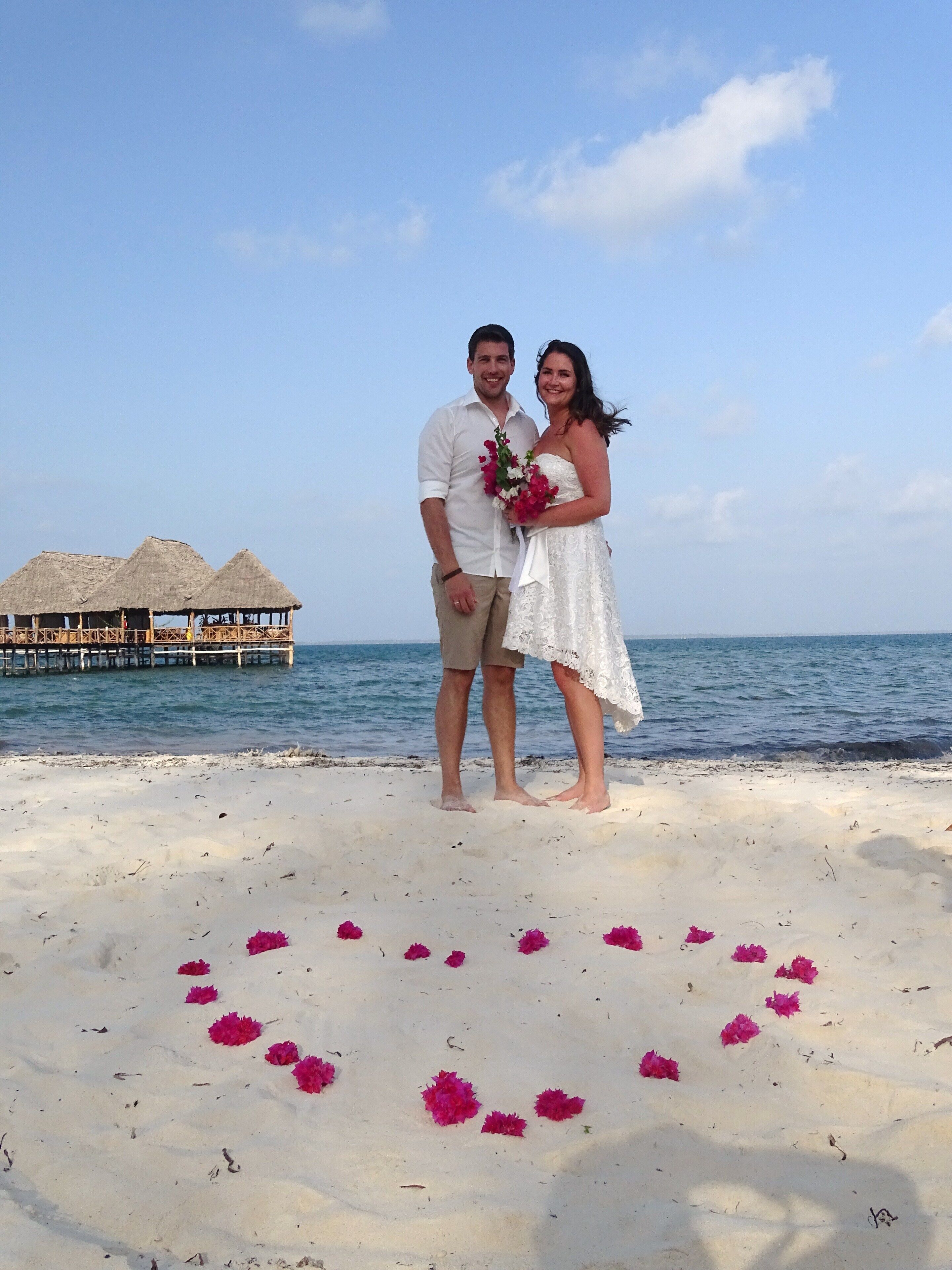 Una pareja parada en la playa, con una estructura de techo de paja al fondo.