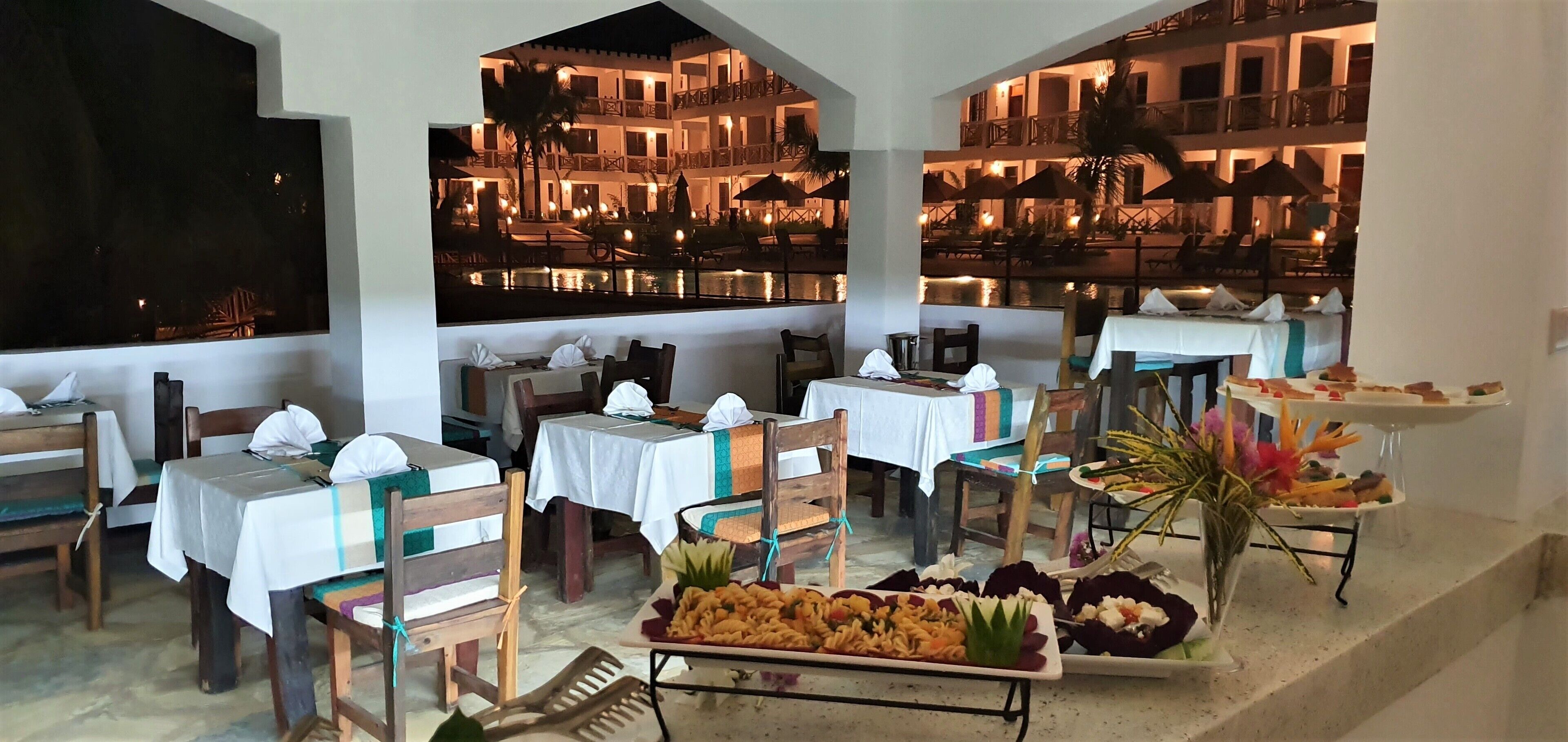Un restaurant avec des tables dressées, un buffet proposant divers plats, et une vue sur un immeuble doté de balcons et d’éclairages.