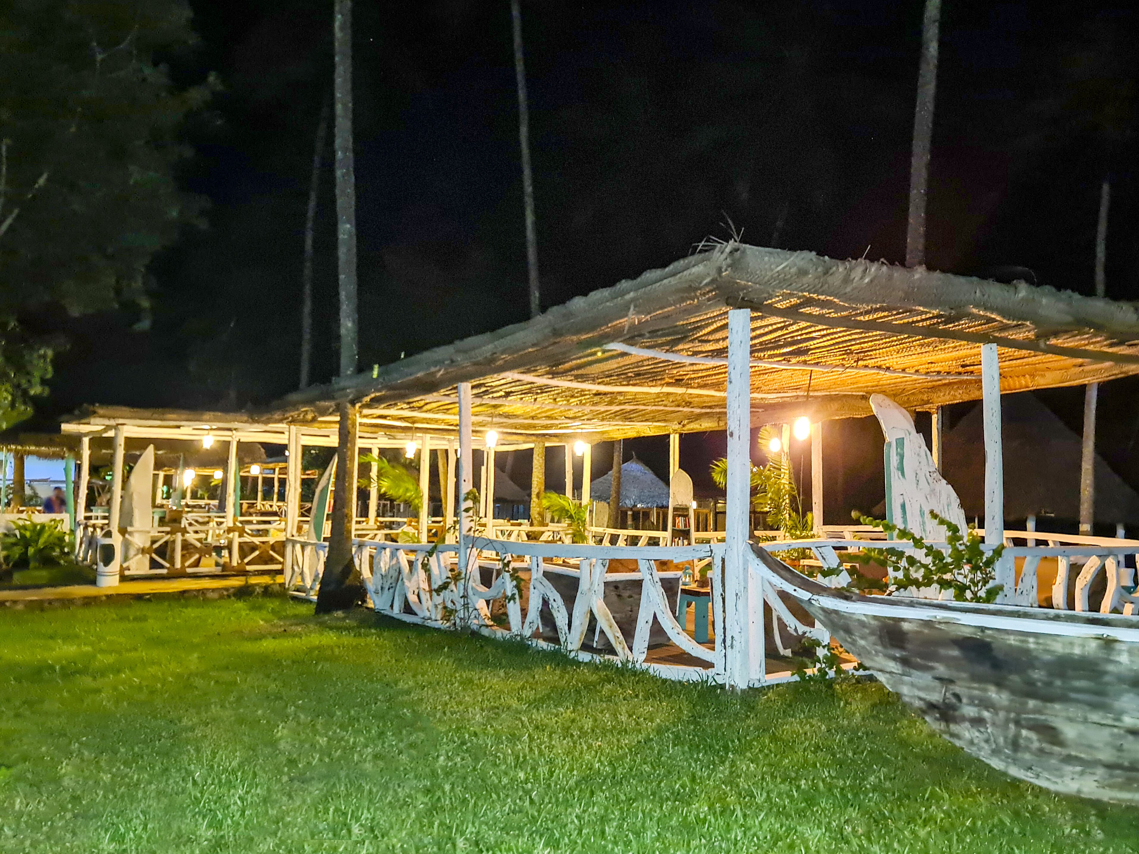 An outdoor seating area with a thatched roof and white railings, surrounded by greenery.