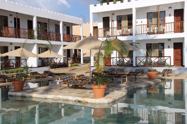 A pool area with lounge chairs and umbrellas, surrounded by multi-story buildings with balconies. - Zanzibar Bay Resort (Marumbi)