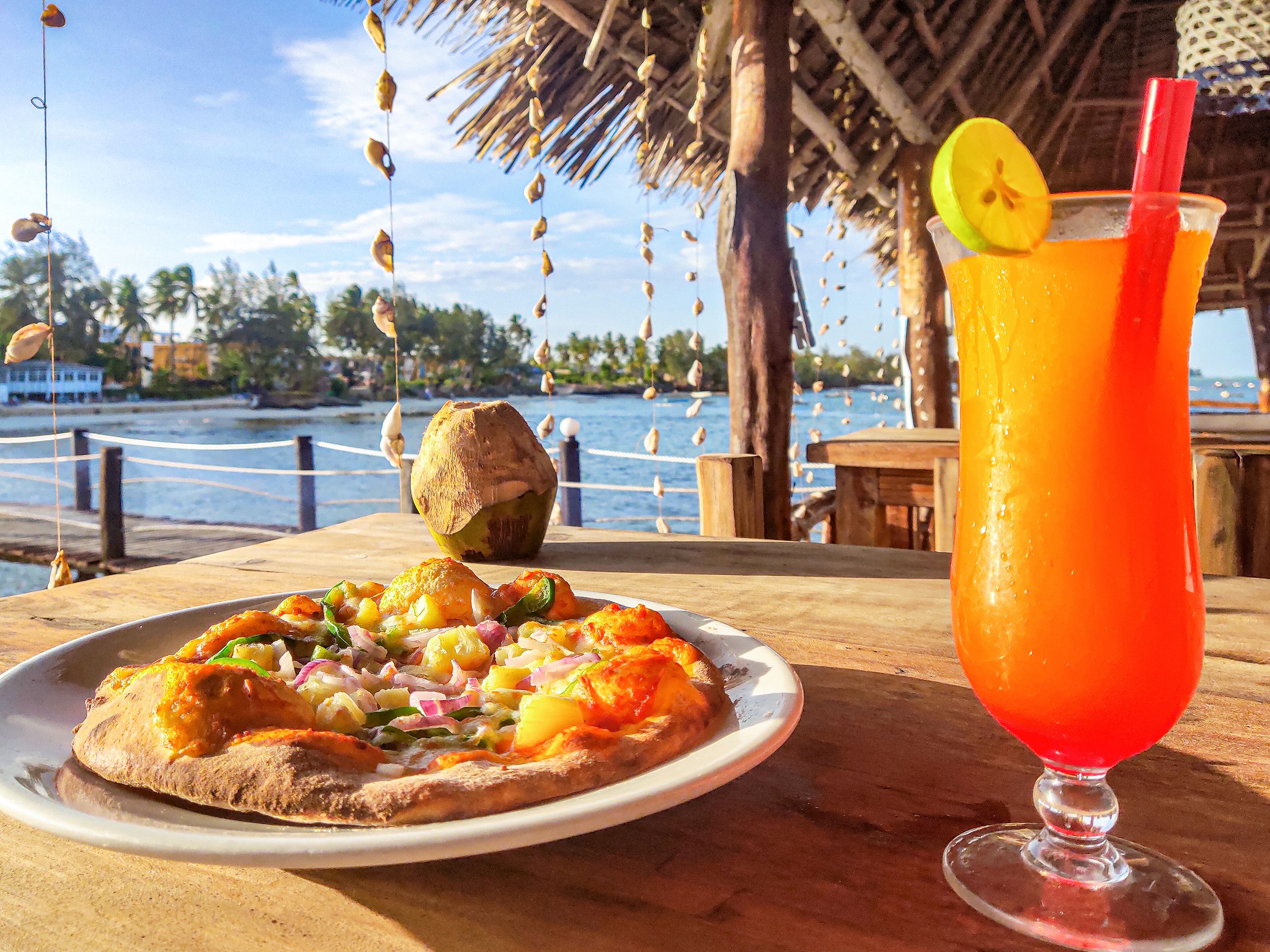 A table with a plate of pizza and a glass of orange drink, set against a backdrop of a beach and palm trees.