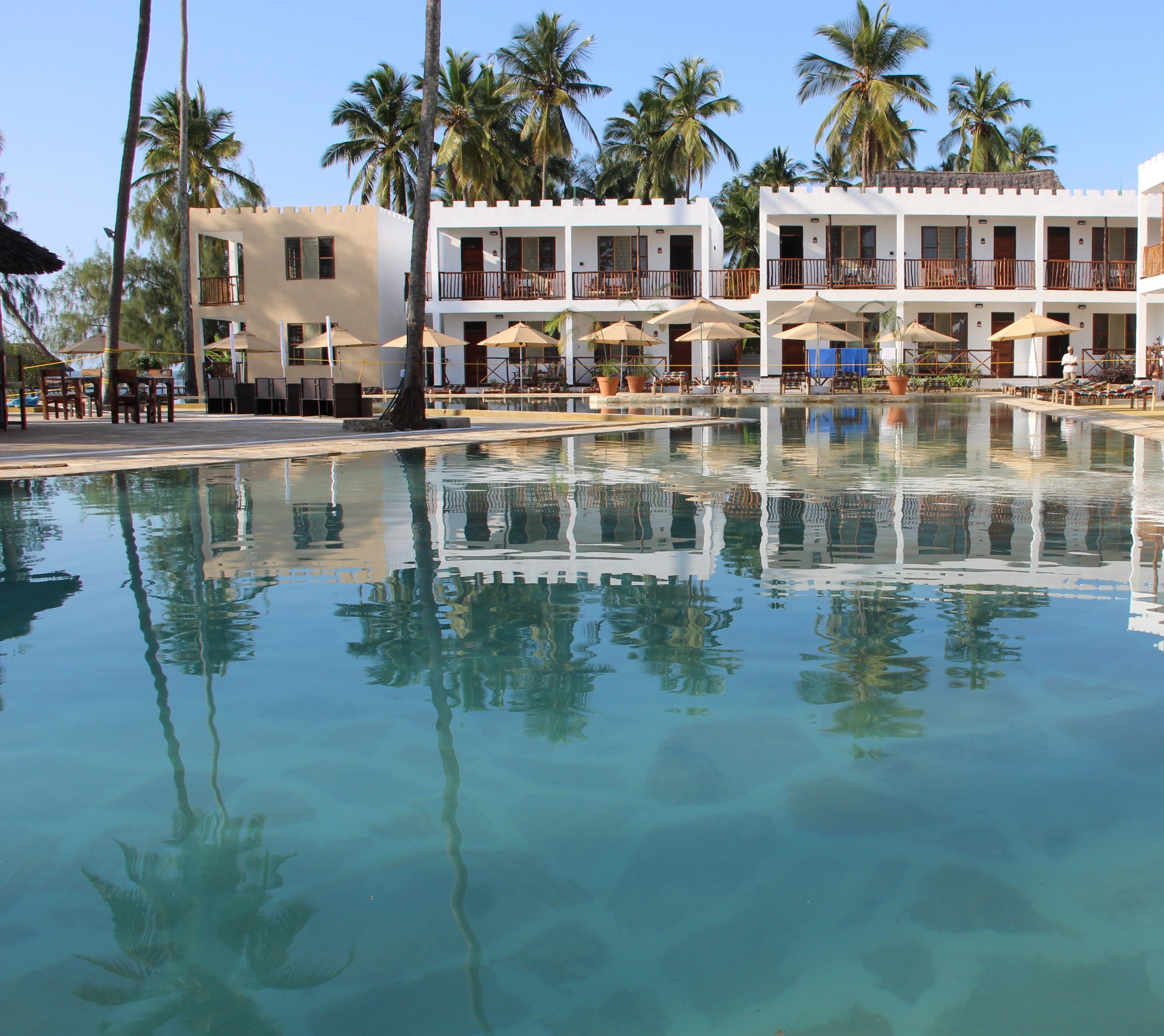 A pool with clear water reflecting palm trees and a building with multiple balconies and outdoor seating.