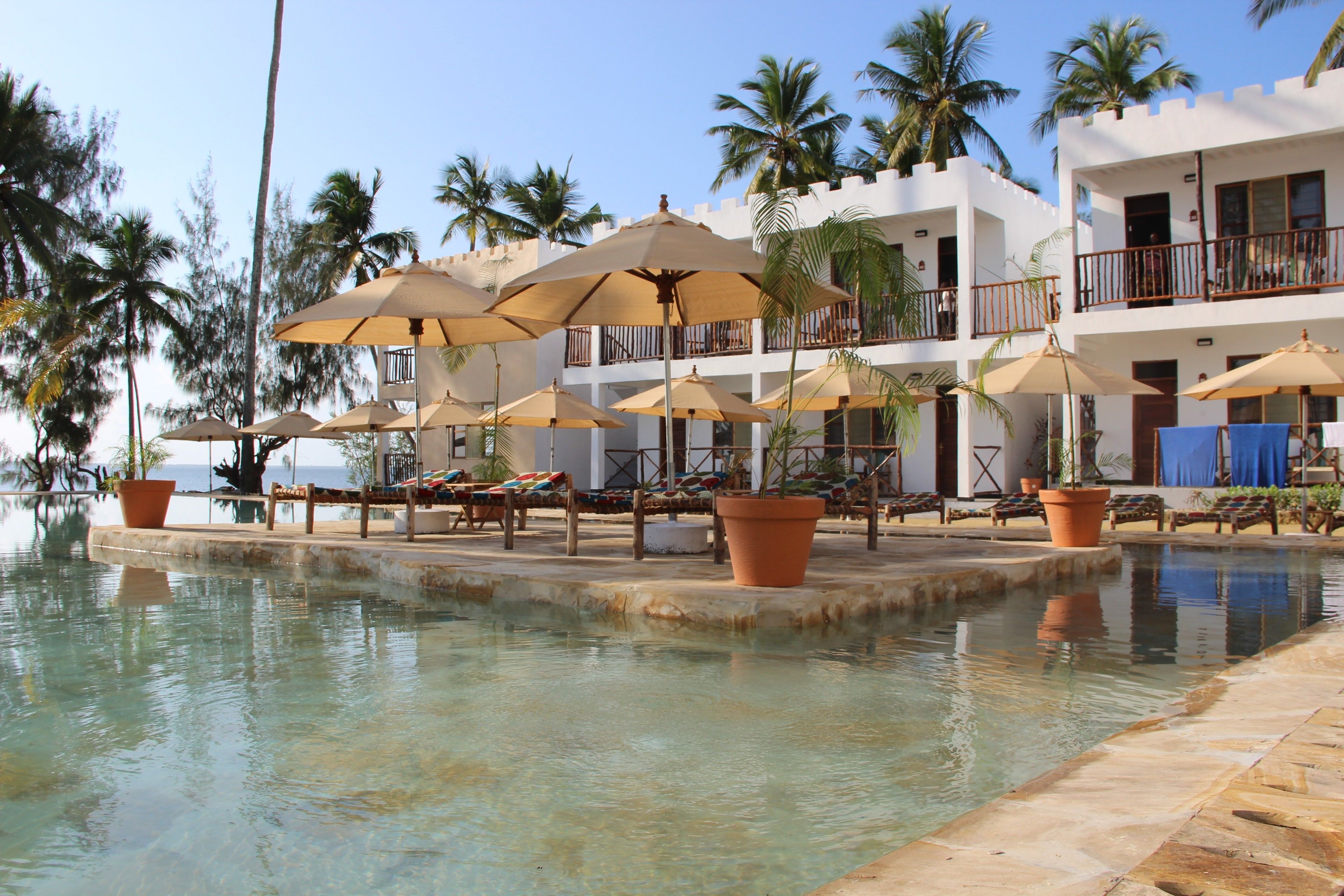 A poolside area with lounge chairs and umbrellas, palm trees, and a building with balconies.