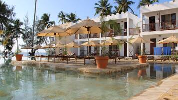 A poolside area with lounge chairs and umbrellas, palm trees, and a building with balconies.