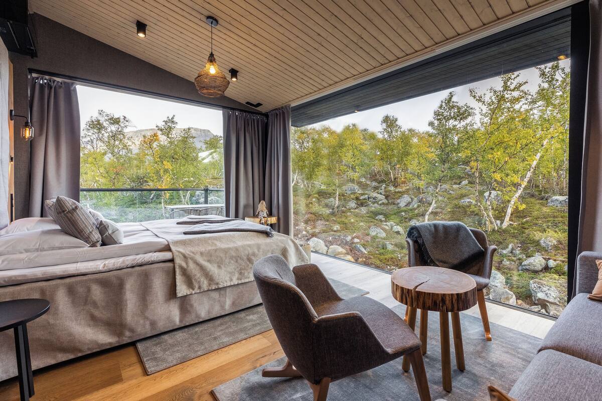 Une chambre moderne avec un grand lit, deux fauteuils, une petite table et une vue sur un espace extérieur rocheux, grâce à de grandes fenêtres.