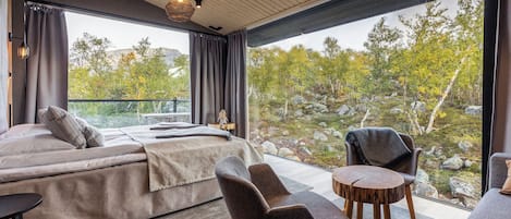 Un dormitorio moderno con una cama grande, dos sillones, una mesita y vistas a un terreno rocoso a través de amplios ventanales.