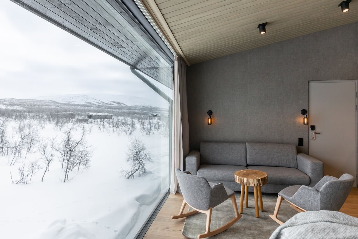 Un salon moderne avec une grande fenêtre, un canapé gris, des fauteuils à bascule en bois et une petite table ronde.