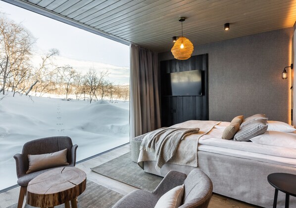 A modern bedroom with a large window, a bed, a TV, and a cozy seating area.