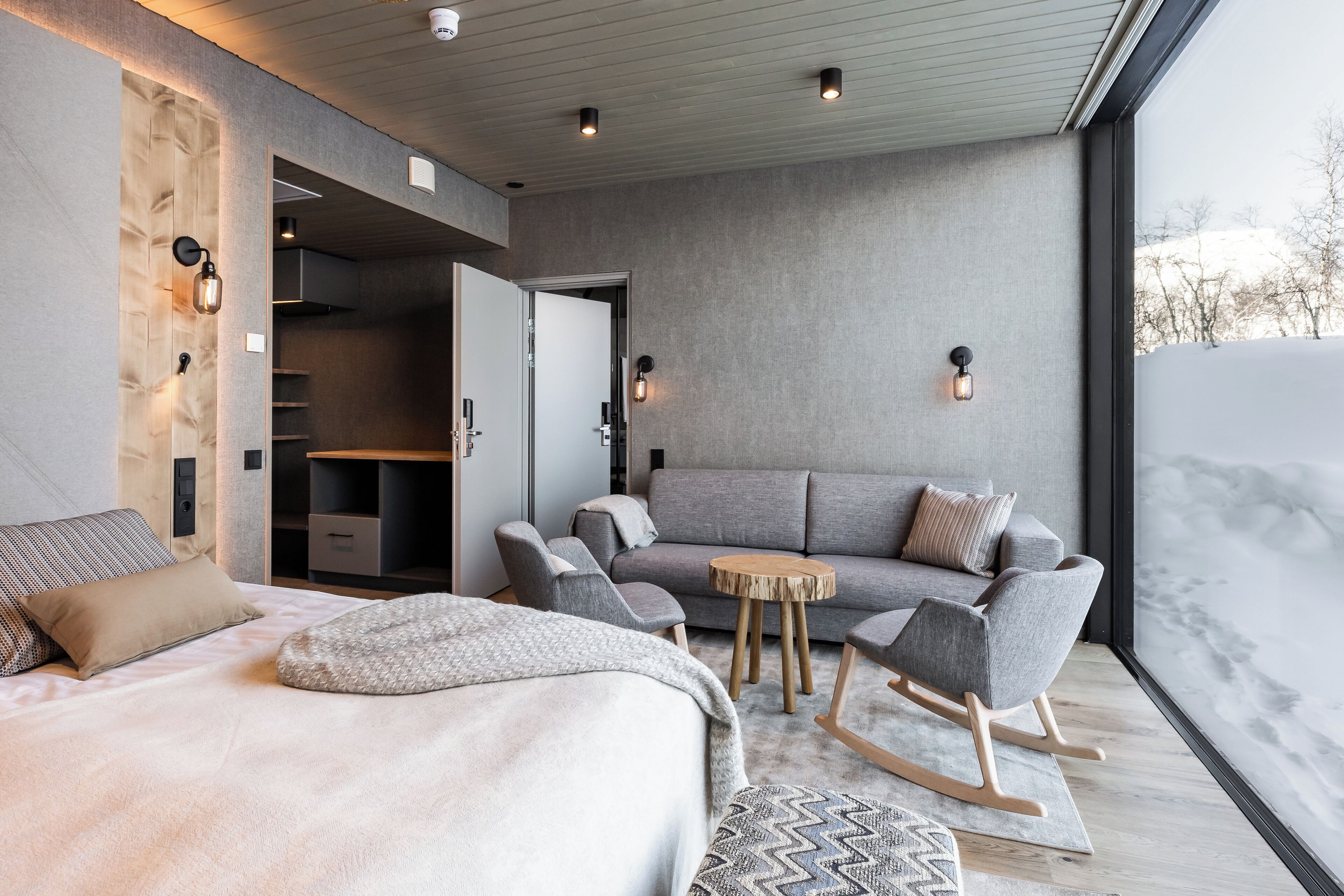 Ein modernes Schlafzimmer mit einem großen Bett, einer Couch und einem Schaukelstuhl. Ein Kleiderschrank mit Schiebetür und ein großer Fensterblick auf eine verschneite Landschaft.