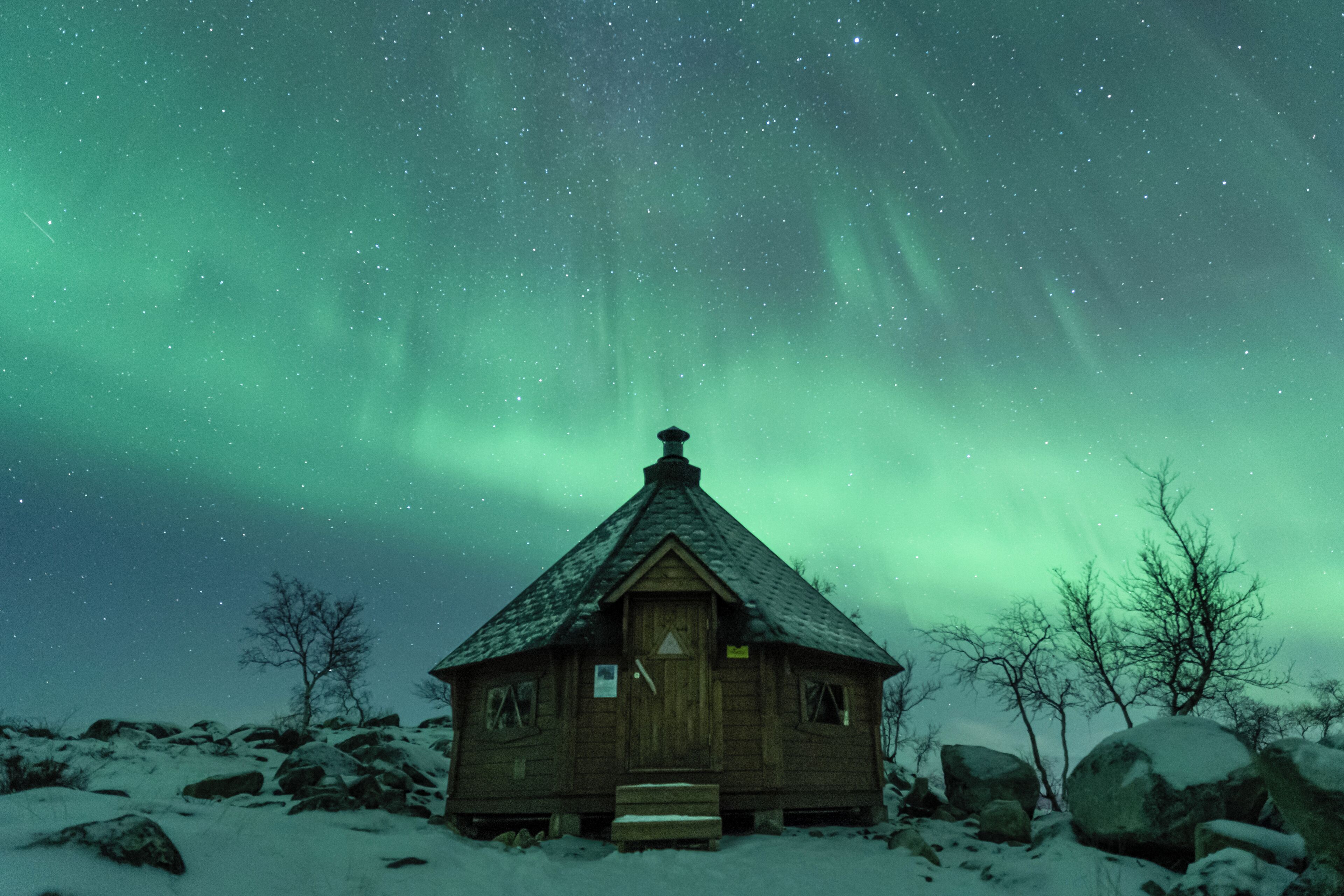 Una cabaña de madera con techo nevado, rodeada de rocas y árboles, bajo un cielo nocturno estrellado.