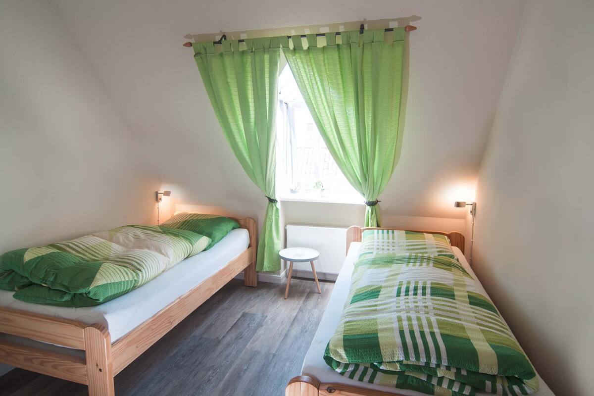 Une petite pièce bien éclairée, avec deux lits en bois, un petit tabouret et une fenêtre ornée de rideaux verts.