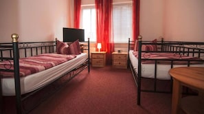 Ein Schlafzimmer mit zwei Einzelbetten, einem kleinen Holztisch, einem Nachttisch mit Lampe und einem an der Wand befestigten Fernseher.