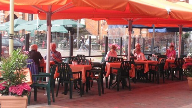 Un café en plein air avec des tables et des chaises sous des parasols orange, entouré de plantes et de fleurs en pot.