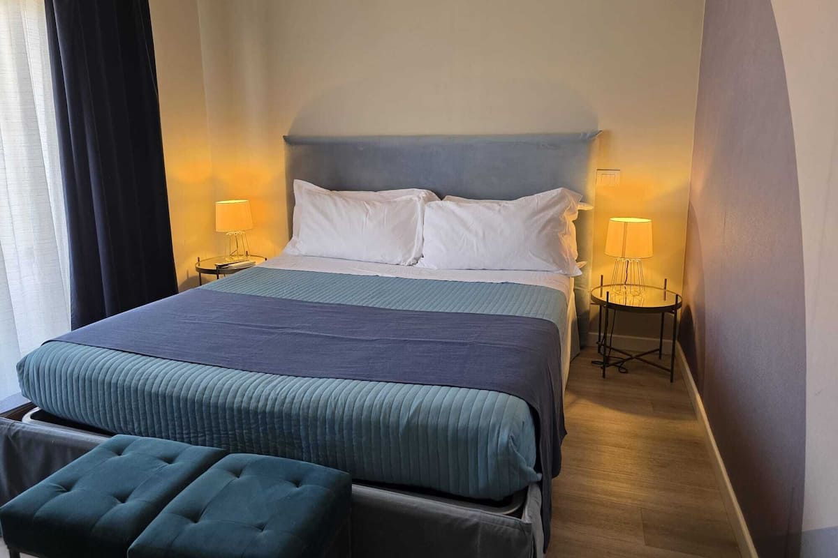Habitación de hotel con una cama grande, dos mesitas de noche con lámparas, un banco al pie de la cama y una ventana con cortinas.