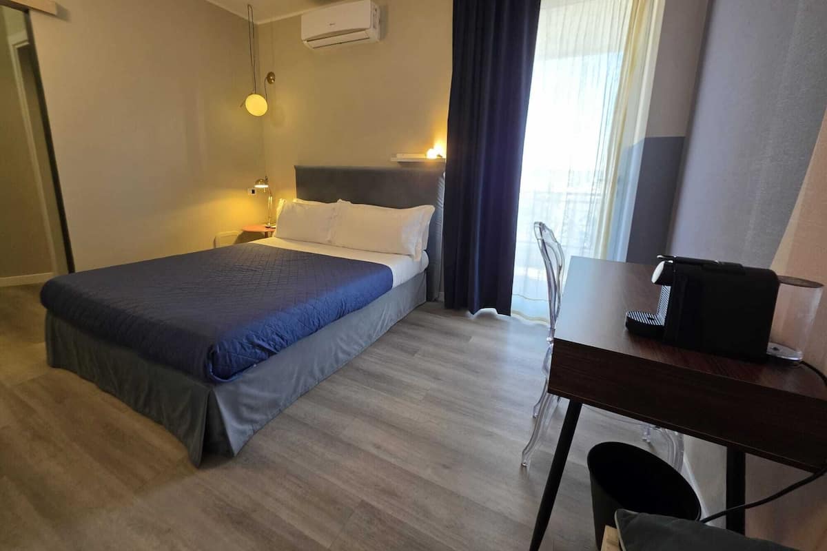 Habitación de hotel con cama, mesita de noche, lámpara, escritorio con silla y ventana con cortinas.