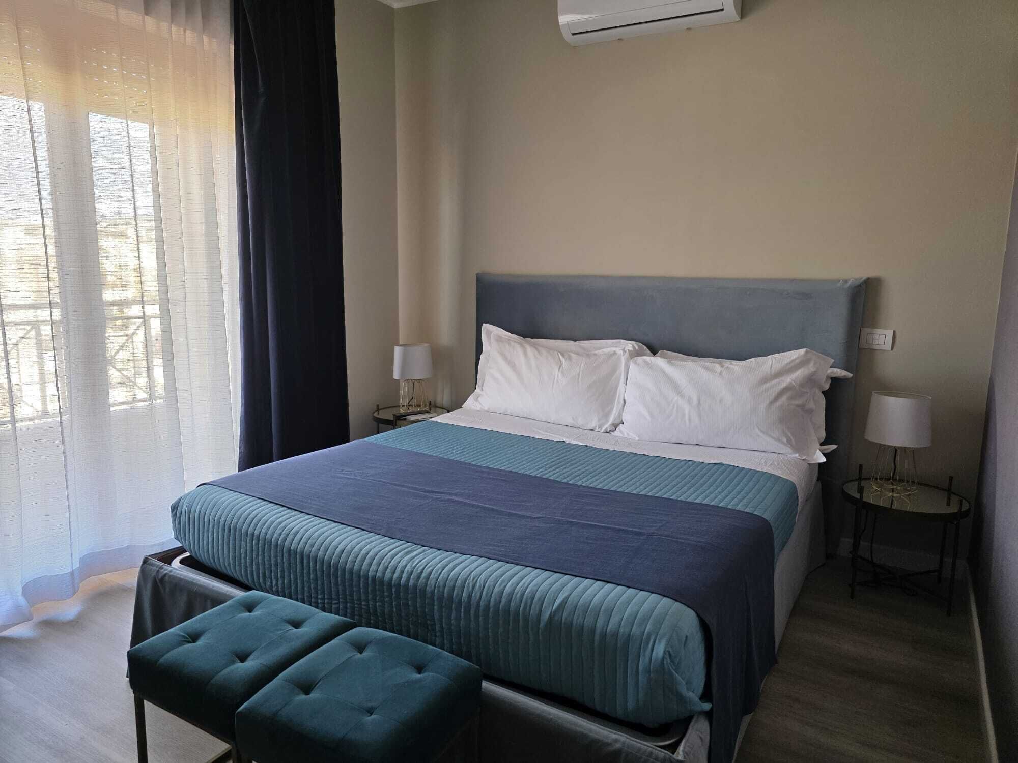 Habitación de hotel con cama, banco, dos mesitas de noche con lámparas y una ventana con cortinas.