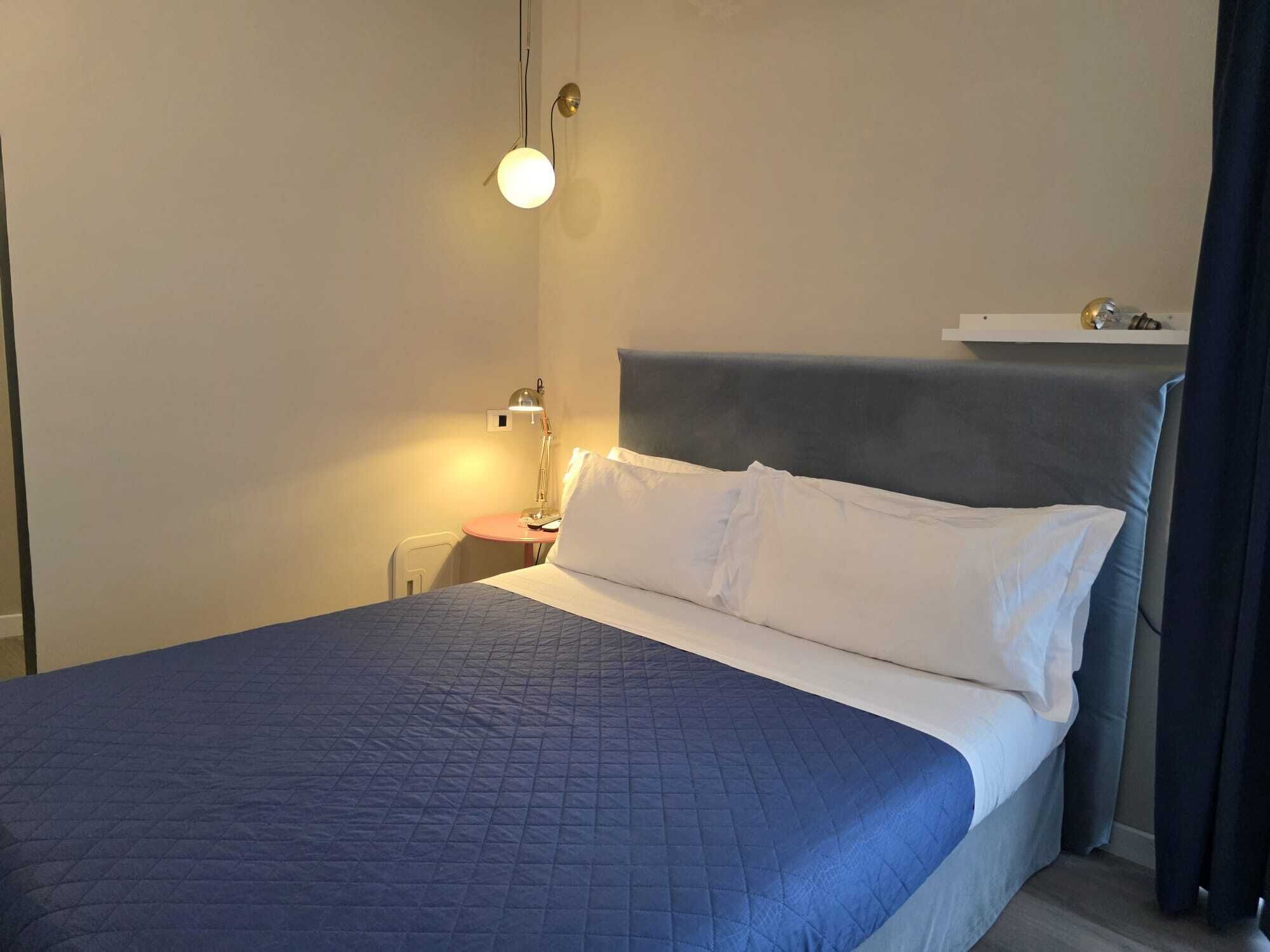 Una cama bien hecha con un edredón azul, una lámpara de noche y una mesita con lámpara.
