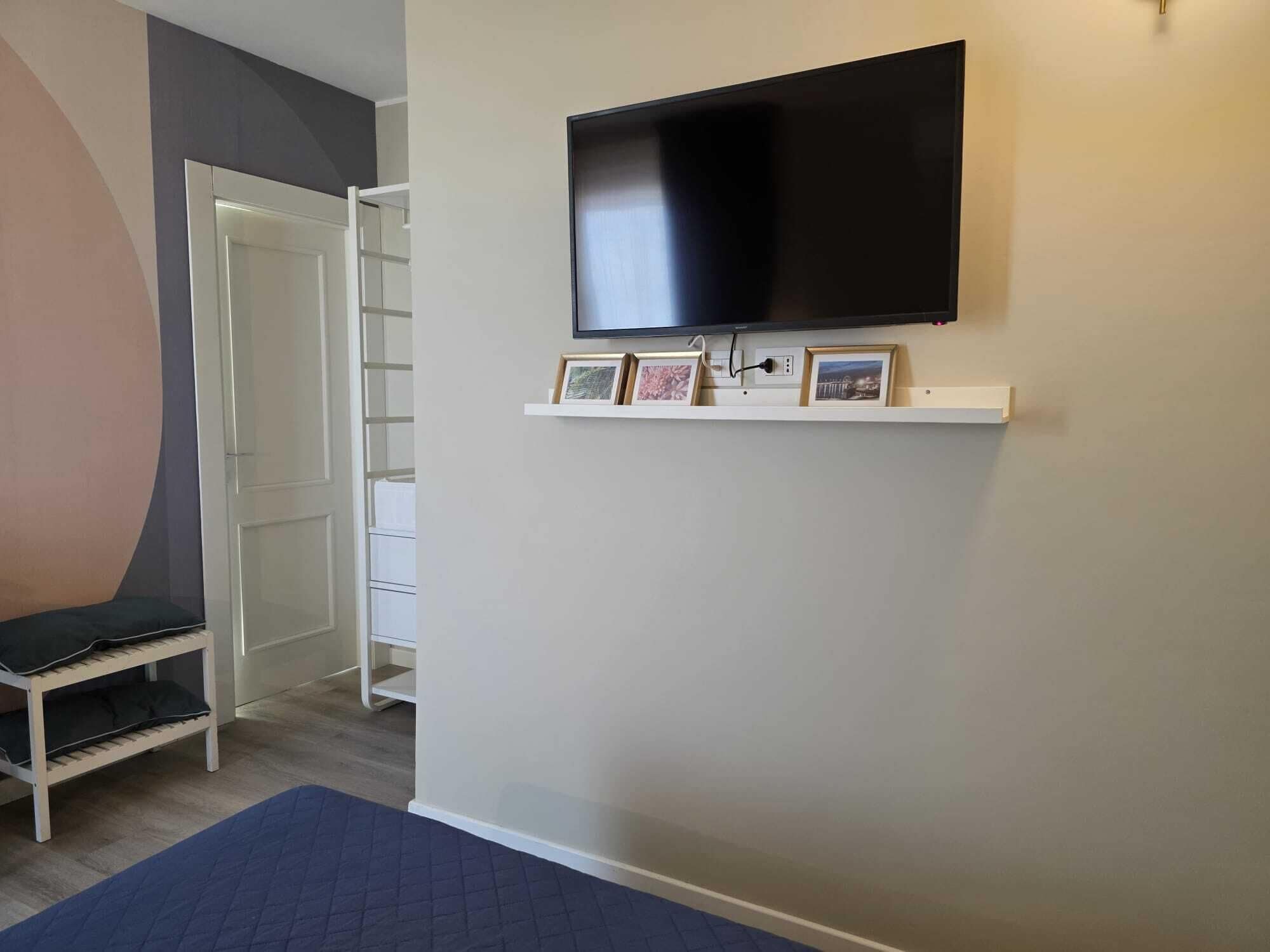 A room with a TV mounted on the wall, a small shelf with pictures, a chair, and a bed.