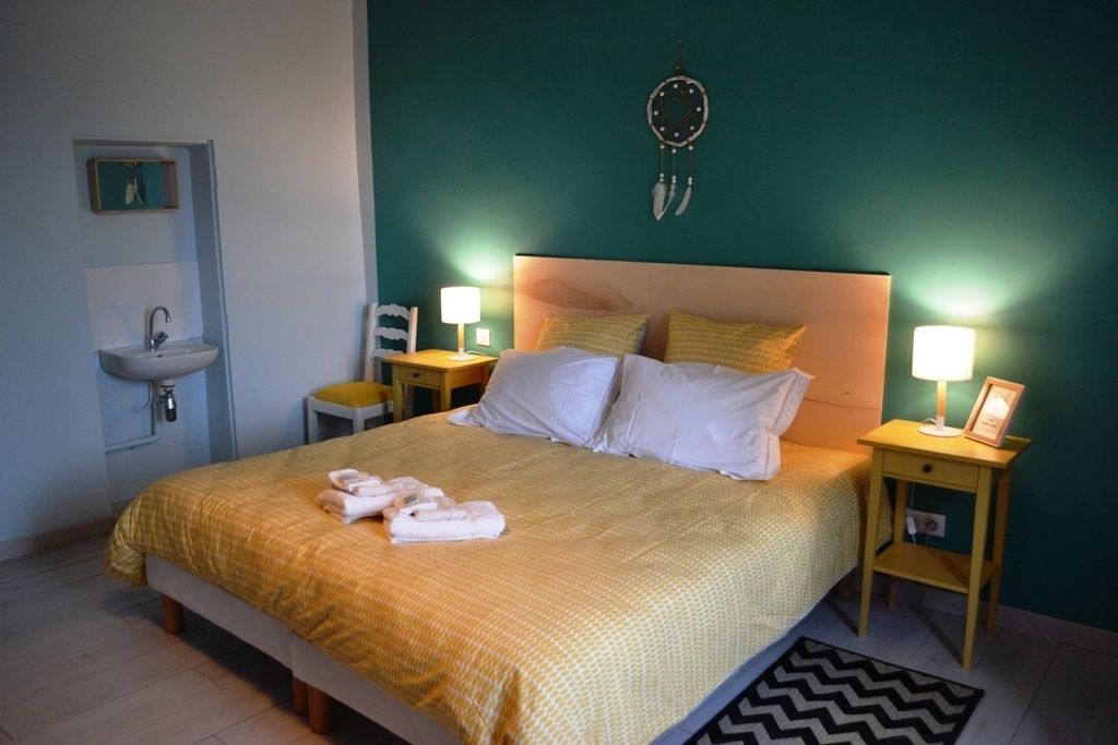 Un lit bien fait, recouvert d’une couvre-lit jaune, avec deux oreillers et une paire de serviettes posées sur une table de chevet. Sur cette table se trouve une lampe et une petite étagère. Les murs de la pièce sont de couleur turquoise et un attrape-rêves est accroché au mur.