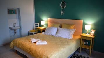 Un lit bien fait, recouvert d’une couvre-lit jaune, avec deux oreillers et une paire de serviettes posées sur une table de chevet. Sur cette table se trouve une lampe et une petite étagère. Les murs de la pièce sont de couleur turquoise et un attrape-rêves est accroché au mur.