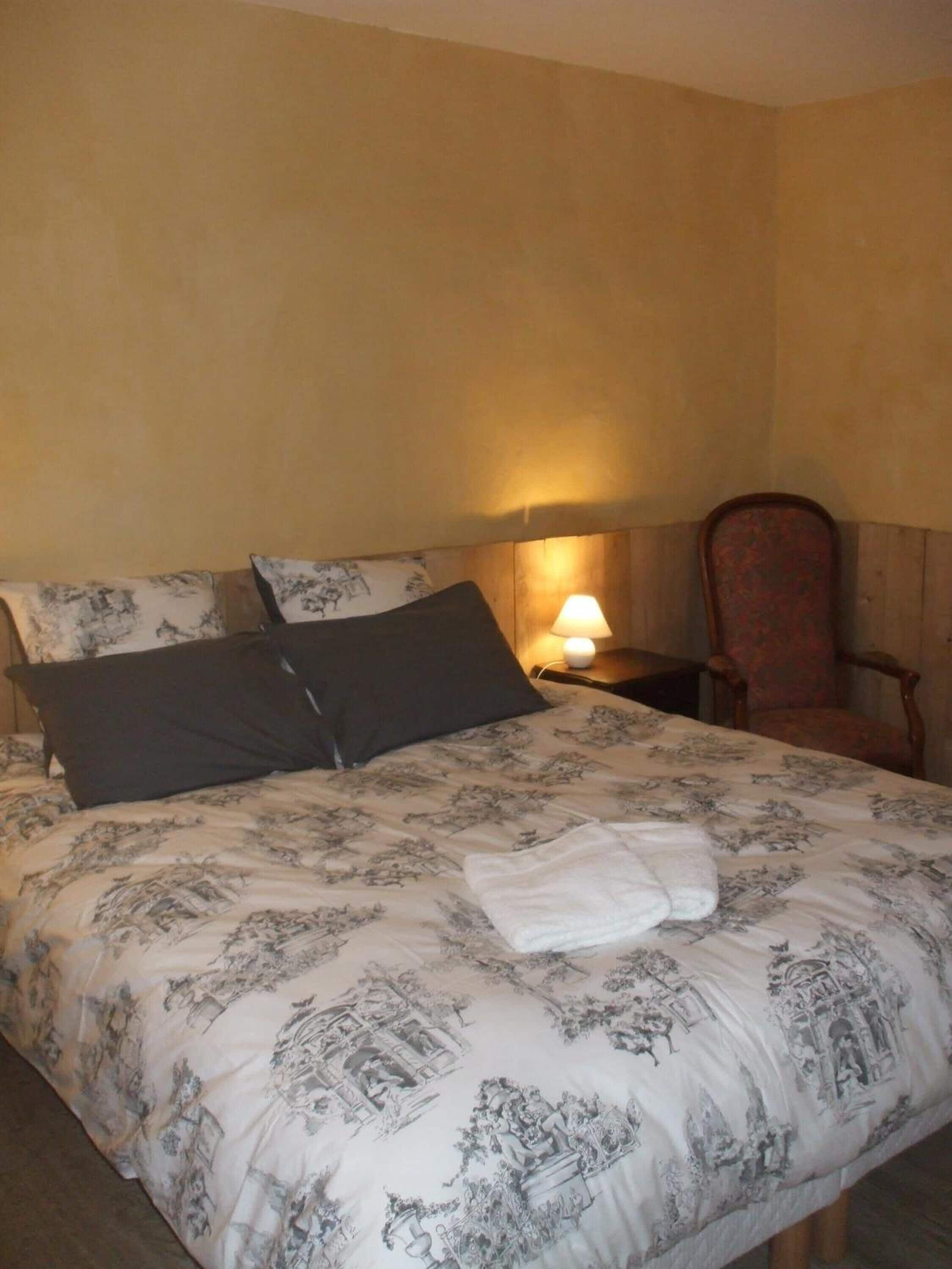 Una cama bien hecha con una colcha de estampado floral, dos almohadas, una toalla doblada, una lámpara de noche y una silla de madera en una habitación con paredes lisas.