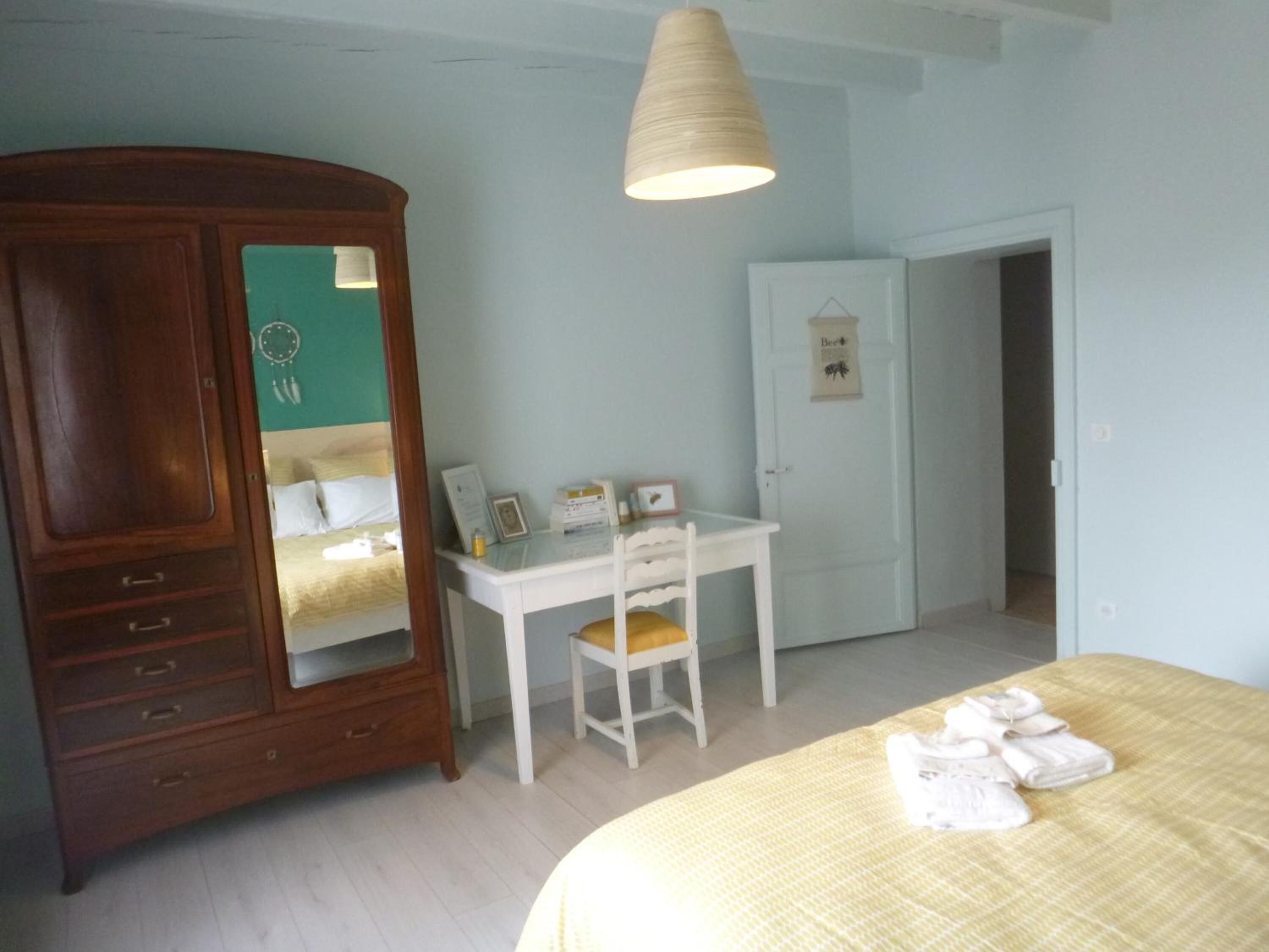 A bedroom with a bed, a wooden wardrobe, a desk with a chair, and a door with a sign.