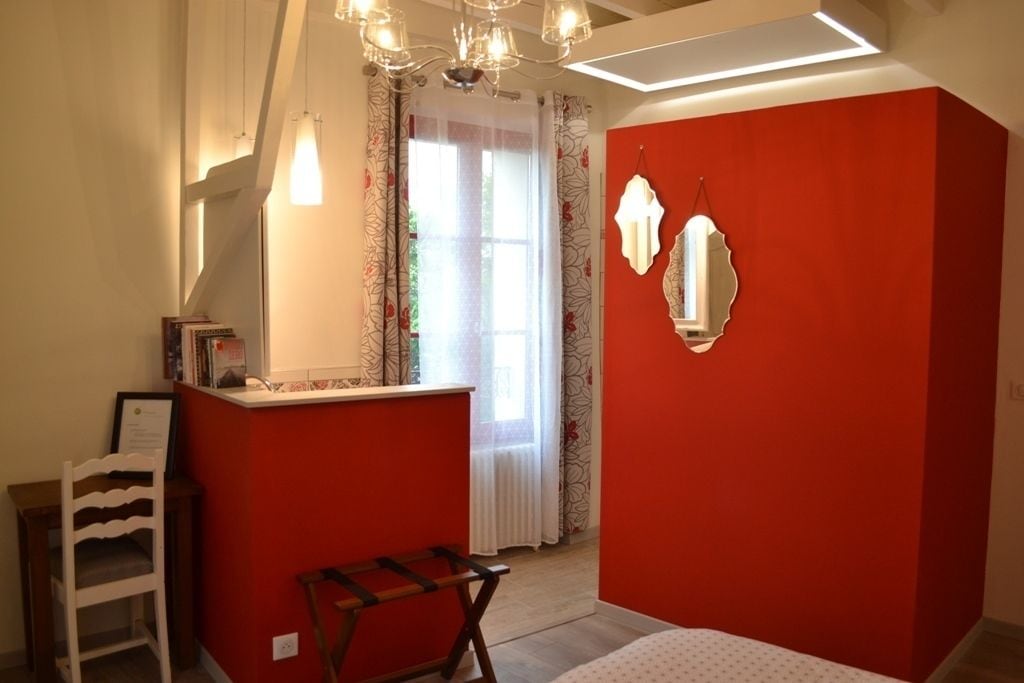 Een slaapkamer met een rode accentmuur, een bed, een bureau met stoel, een hangspiegel en een kroonluchter.
