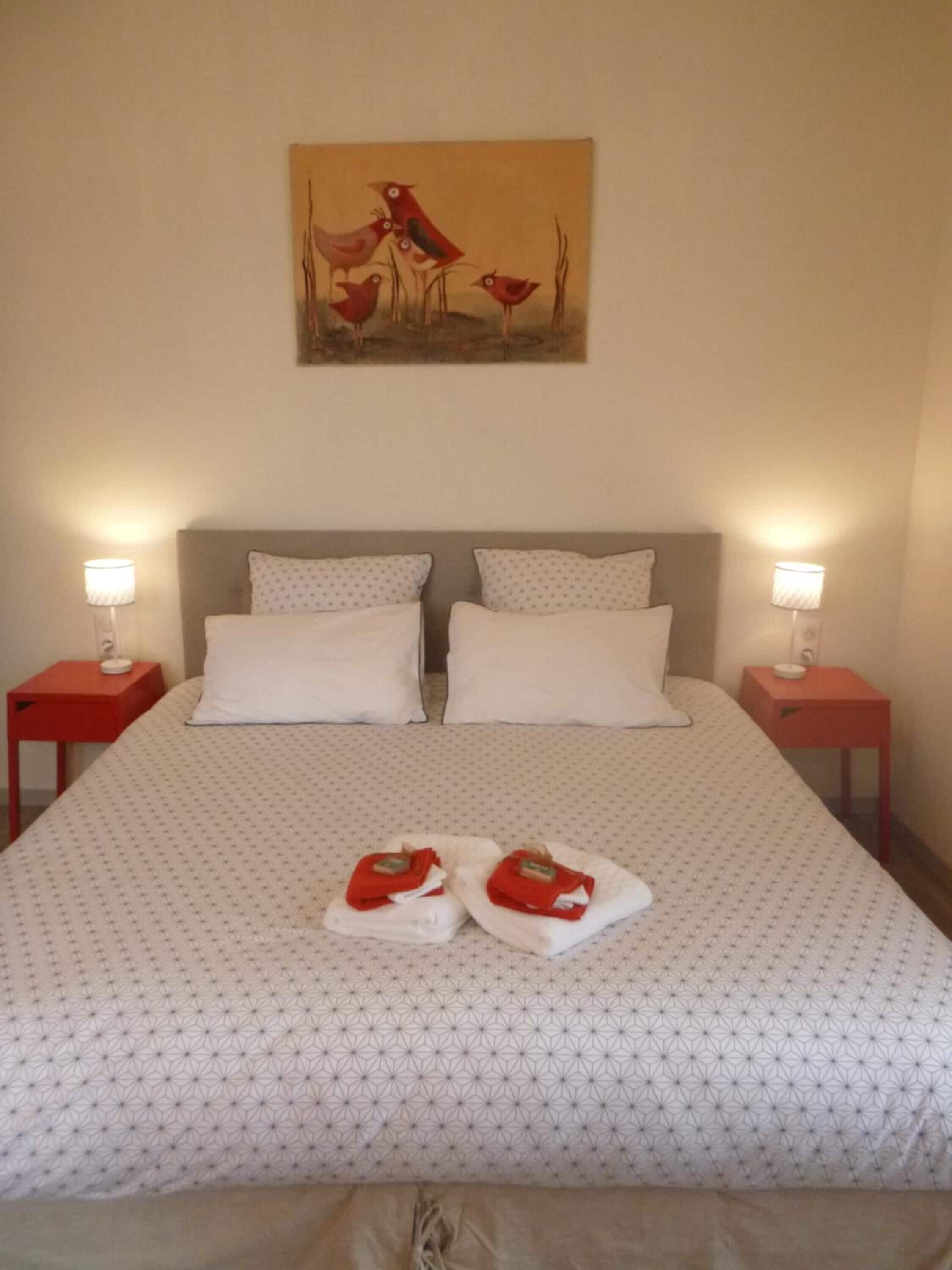 A neatly made bed with a patterned bedspread, two pillows, and two red towels on the bed. There is a painting on the wall above the bed.