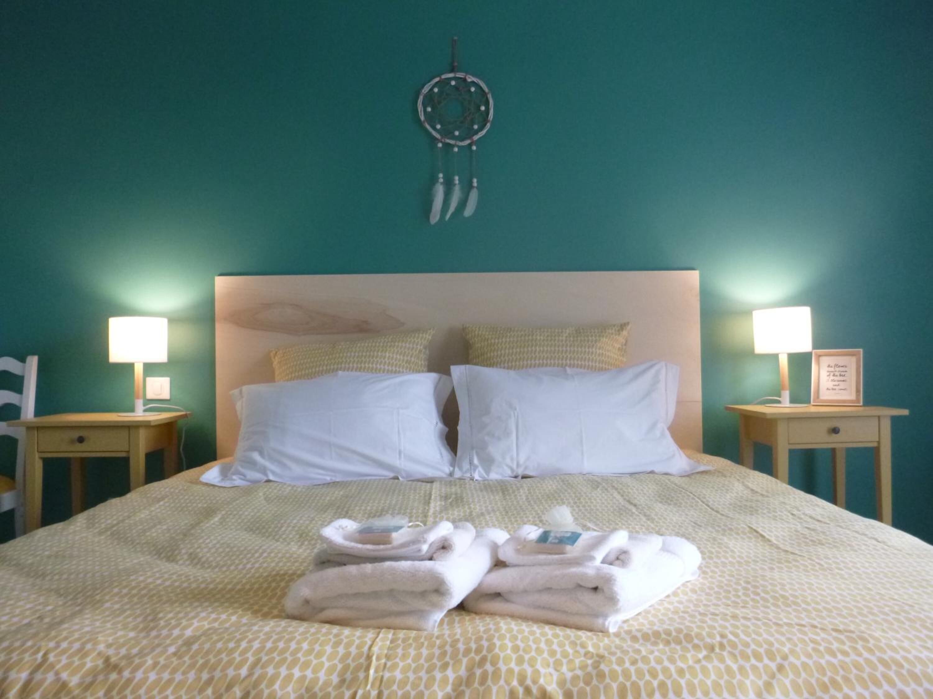 A neatly made bed with white linens, two pillows, and a dreamcatcher hanging above. There are two bedside tables with lamps and a framed picture on one.