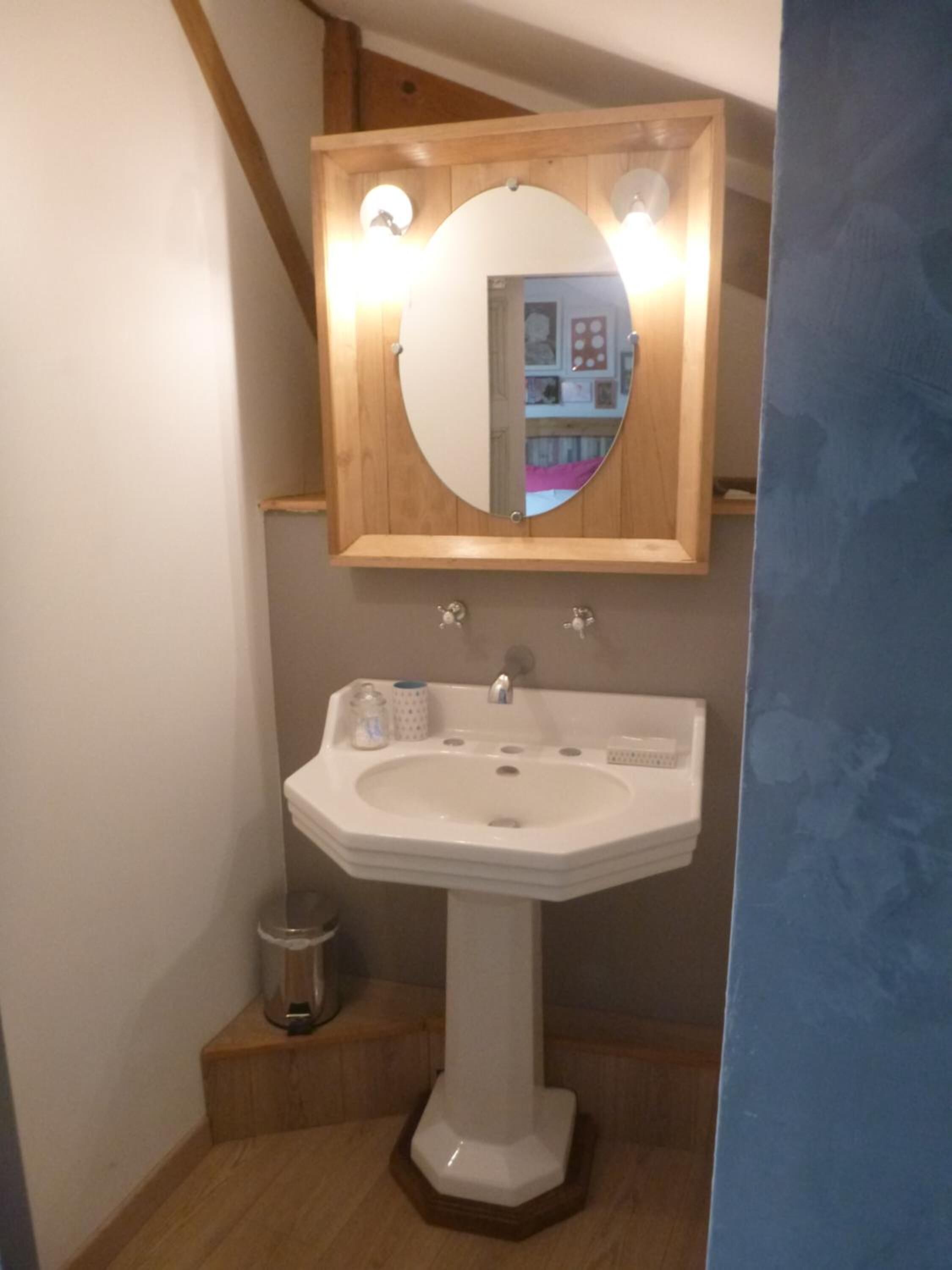 Une salle de bain avec un lavabo sur colonne blanc, un miroir à cadre en bois et une petite poubelle.