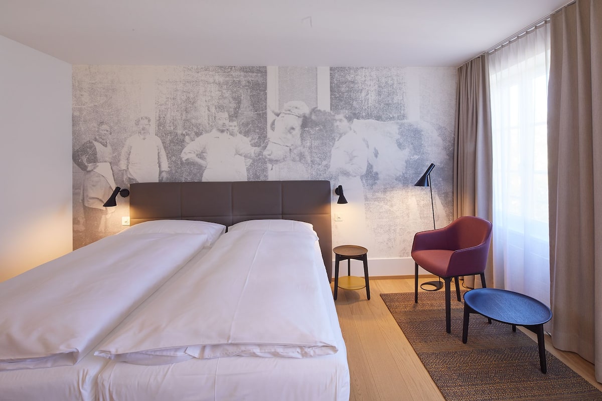 A hotel room with a large bed, a small table, a chair, and a wall-mounted lamp.
