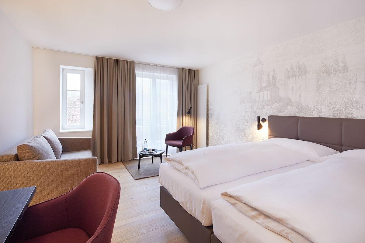 A hotel room with a bed, a sofa, a chair, a small table, and a window with curtains.