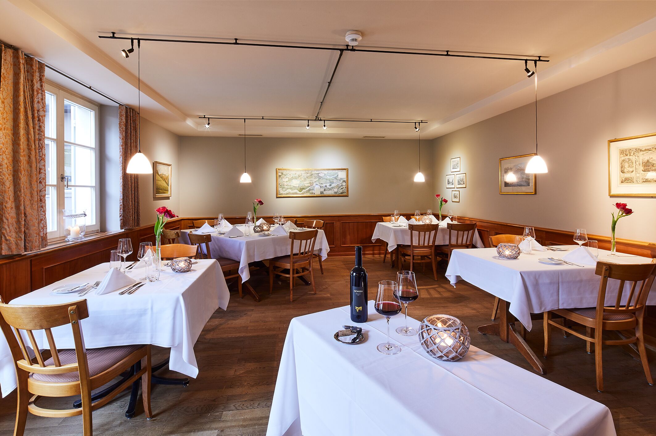 Un comedor con varias mesas dispuestas para comer, cada una con mantel blanco, sillas y copas de vino.
