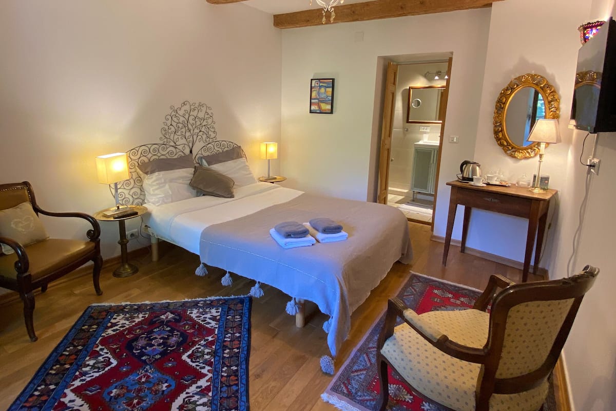 A cozy bedroom with a bed, two chairs, a nightstand, a dresser, a mirror, and a chandelier.