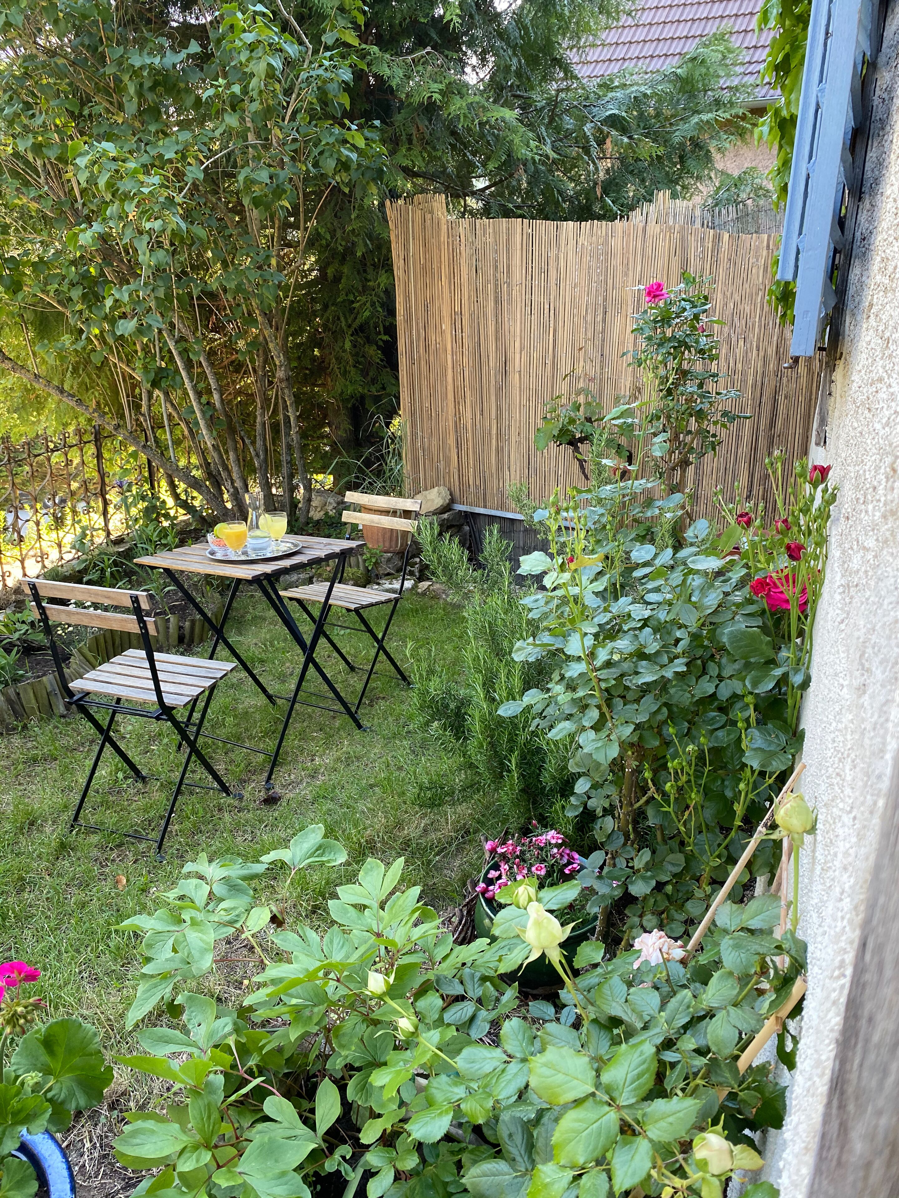 Une petite terrasse extérieure avec une table et des chaises, entourée d’une végétation luxuriante et de plantes en fleurs.