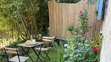 Une petite terrasse extérieure avec une table et des chaises, entourée d’une végétation luxuriante et de plantes en fleurs.