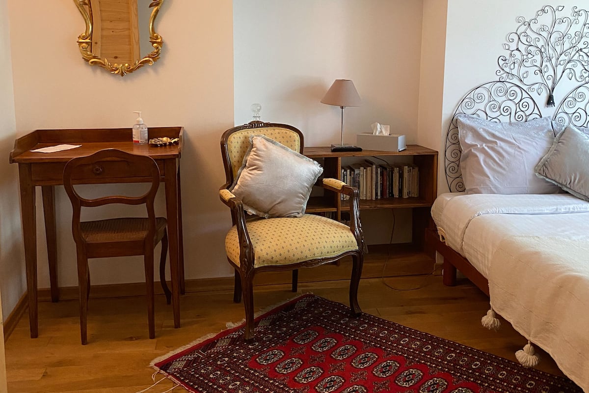 A bedroom with a wooden bed, a chair, a desk, a bookshelf, and a patterned rug.