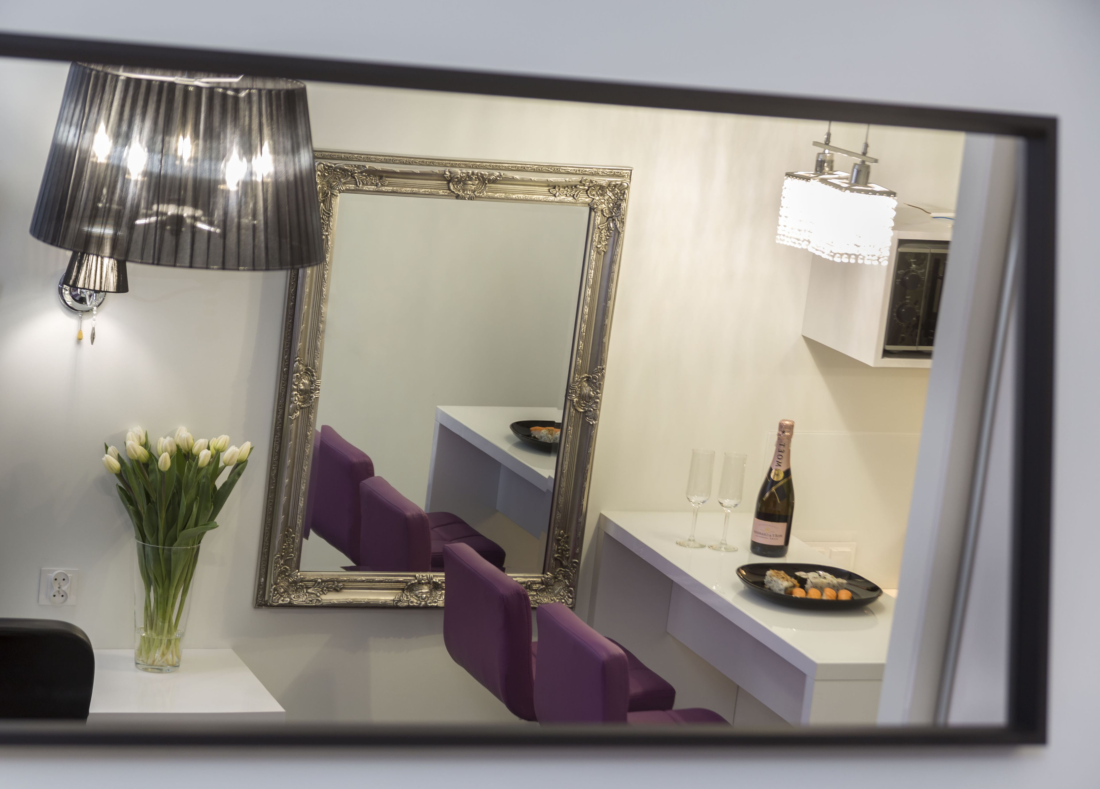 A modern room with a large mirror, purple chairs, a vase of flowers, a bottle of champagne, and a small table with a plate of food.