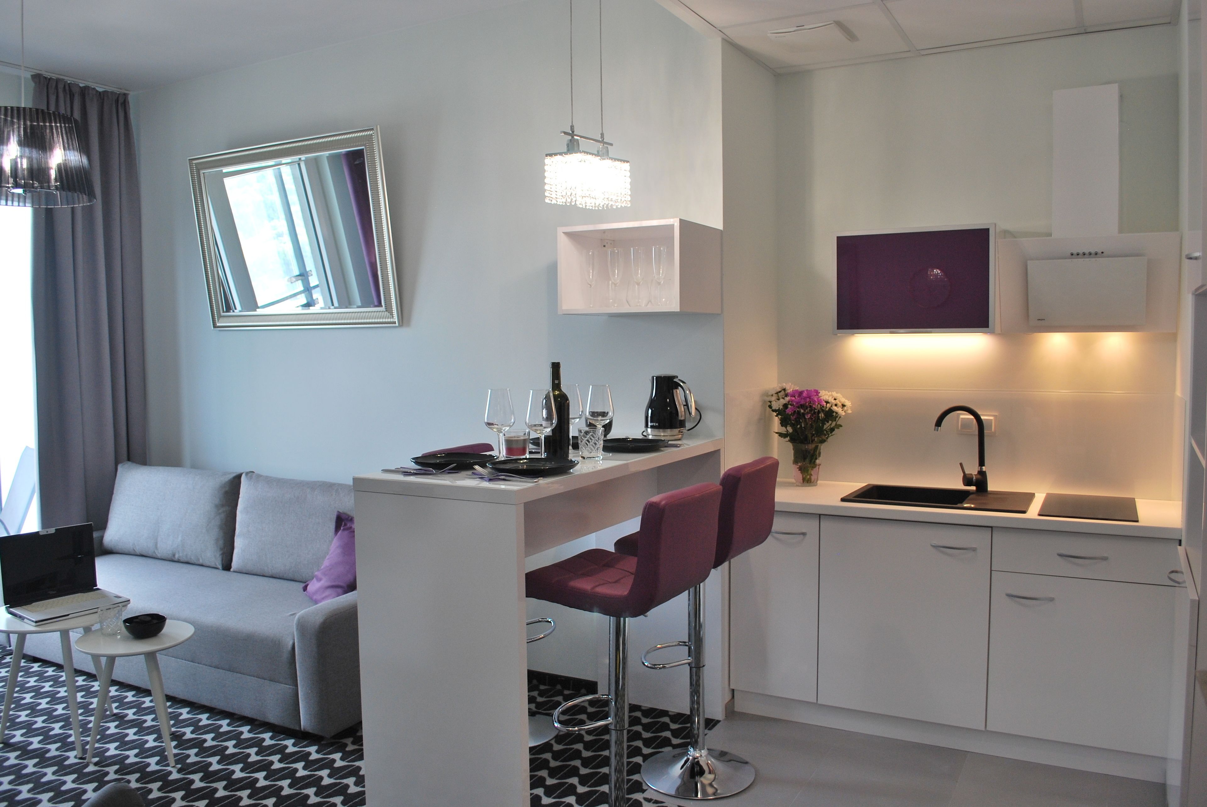 Un espace de vie moderne et compact comprenant un canapé, une petite table à manger, deux tabourets de bar, un coin cuisine avec un évier et des placards, et un miroir au mur.