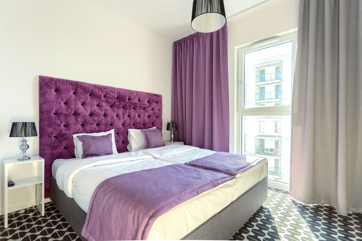 A modern bedroom with a tufted headboard, a large window with curtains, a patterned floor, and a bedside table with a lamp.