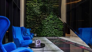 A modern indoor seating area with blue chairs and a small table, a green vertical garden, and a glass railing overlooking a reflective pool. - Vola Residence (Warsaw)