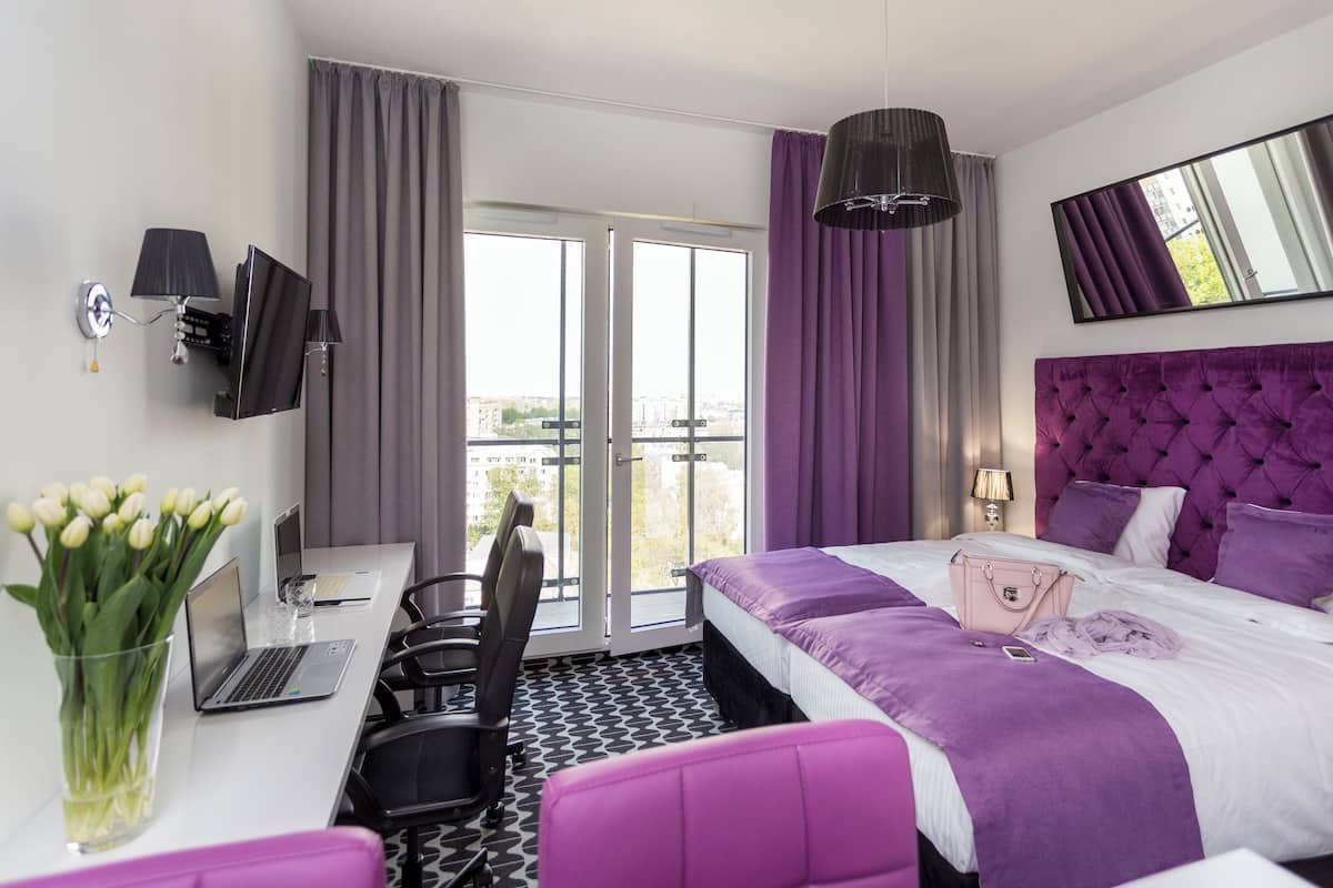 A modern hotel room with a bed, a desk with a laptop, a chair, and a view of the city through large windows.