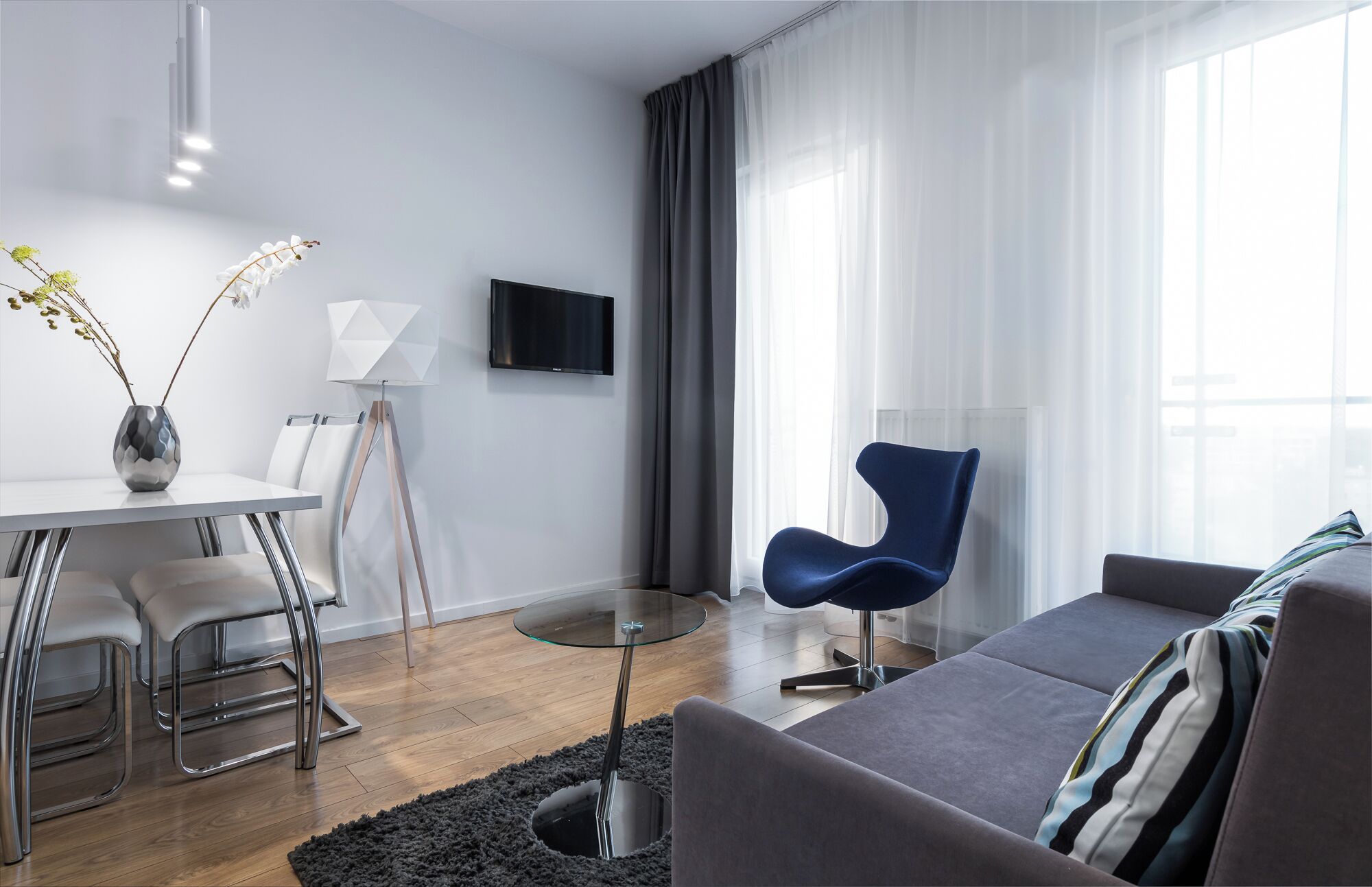 A modern living room with a glass coffee table, a blue chair, a grey sofa, a white TV, and a vase with flowers.