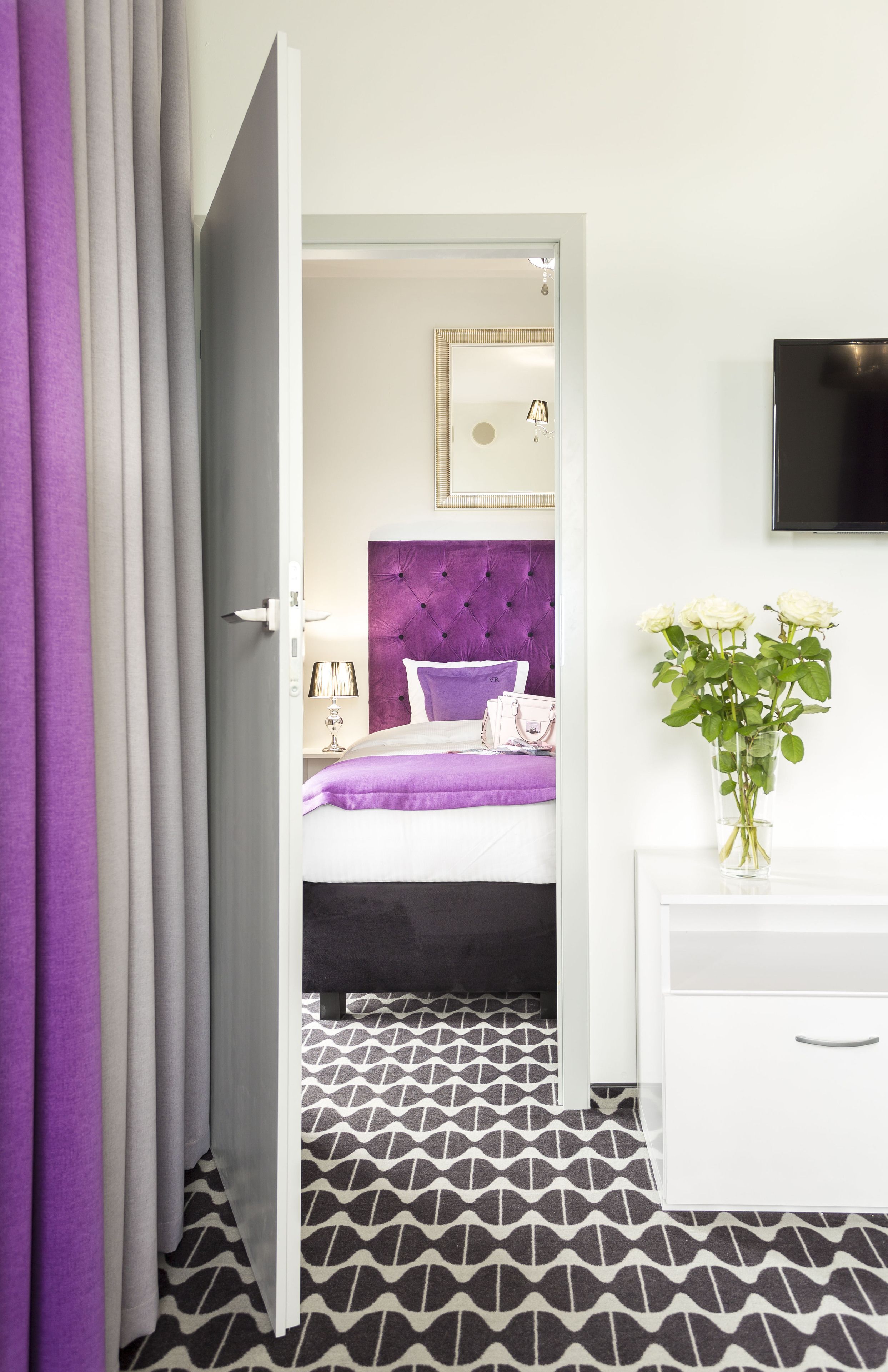 A modern hotel room with a bed, a dresser, a TV, and a vase of flowers.