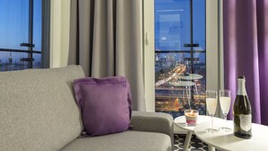A hotel room with a sofa, a small table with glasses and a bottle, and a view of a cityscape through a window. - Vola Residence (Warsaw)