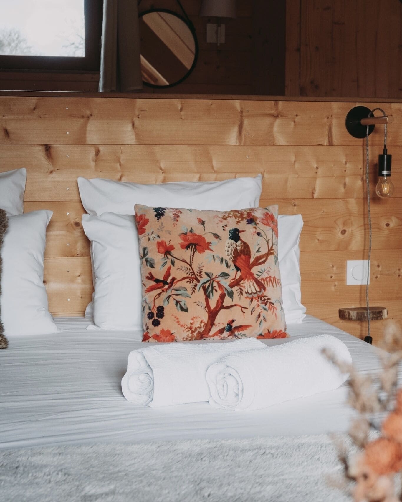A neatly made bed with white linens and a floral pillow, a wooden headboard, and a bedside lamp.