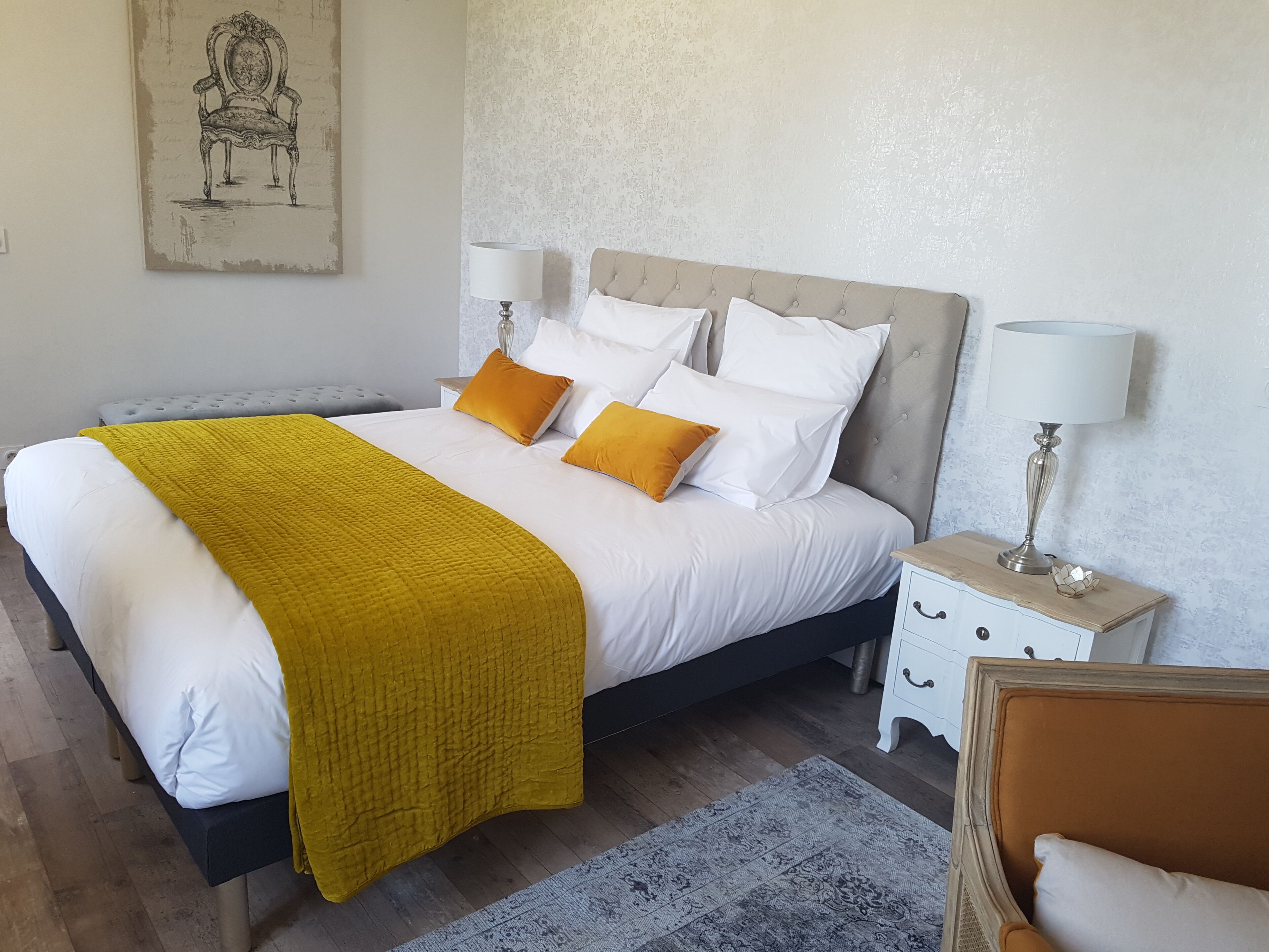 Un lit bien fait, avec une literie blanche et jaune, une tête de lit grise et deux tables de chevet avec des lampes.