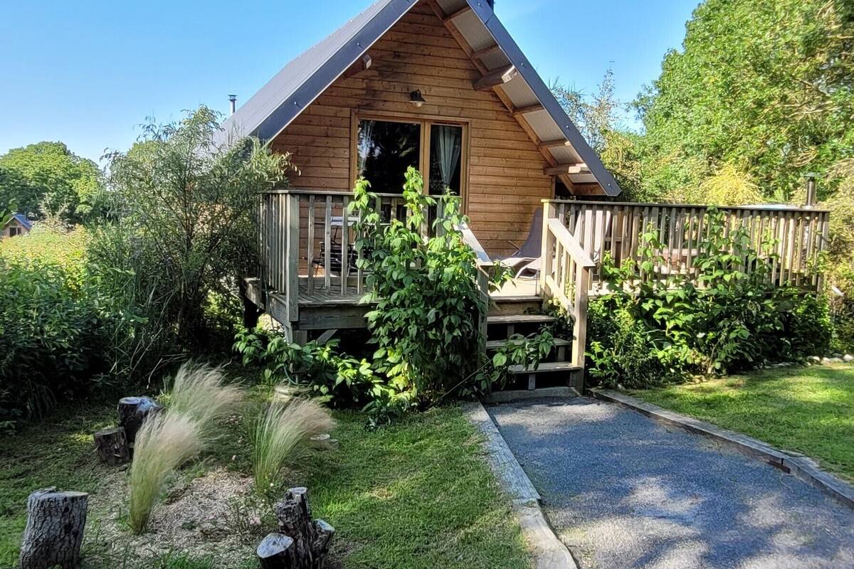 A wooden cabin with a porch, surrounded by lush greenery and a gravel path leading to the entrance.
