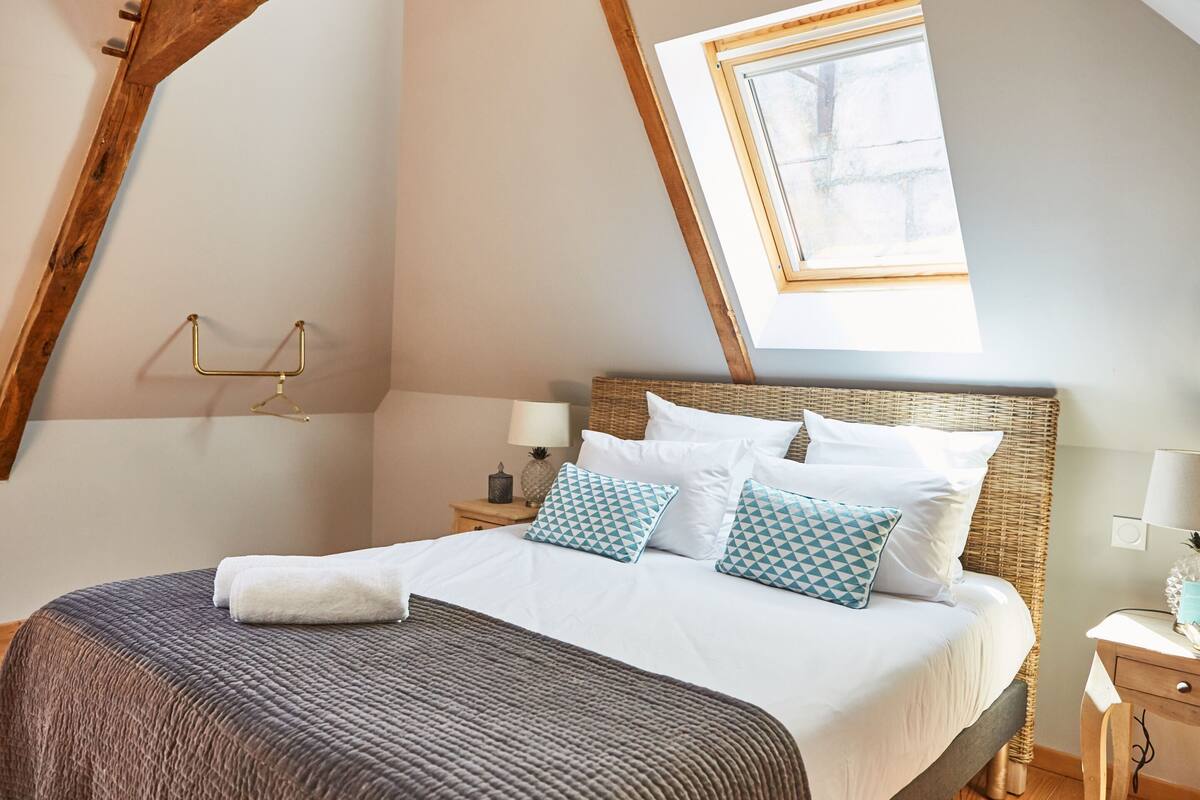 A bedroom with a sloped ceiling, a bed with white bedding and patterned pillows, a bedside table with a lamp, and a small nightstand.